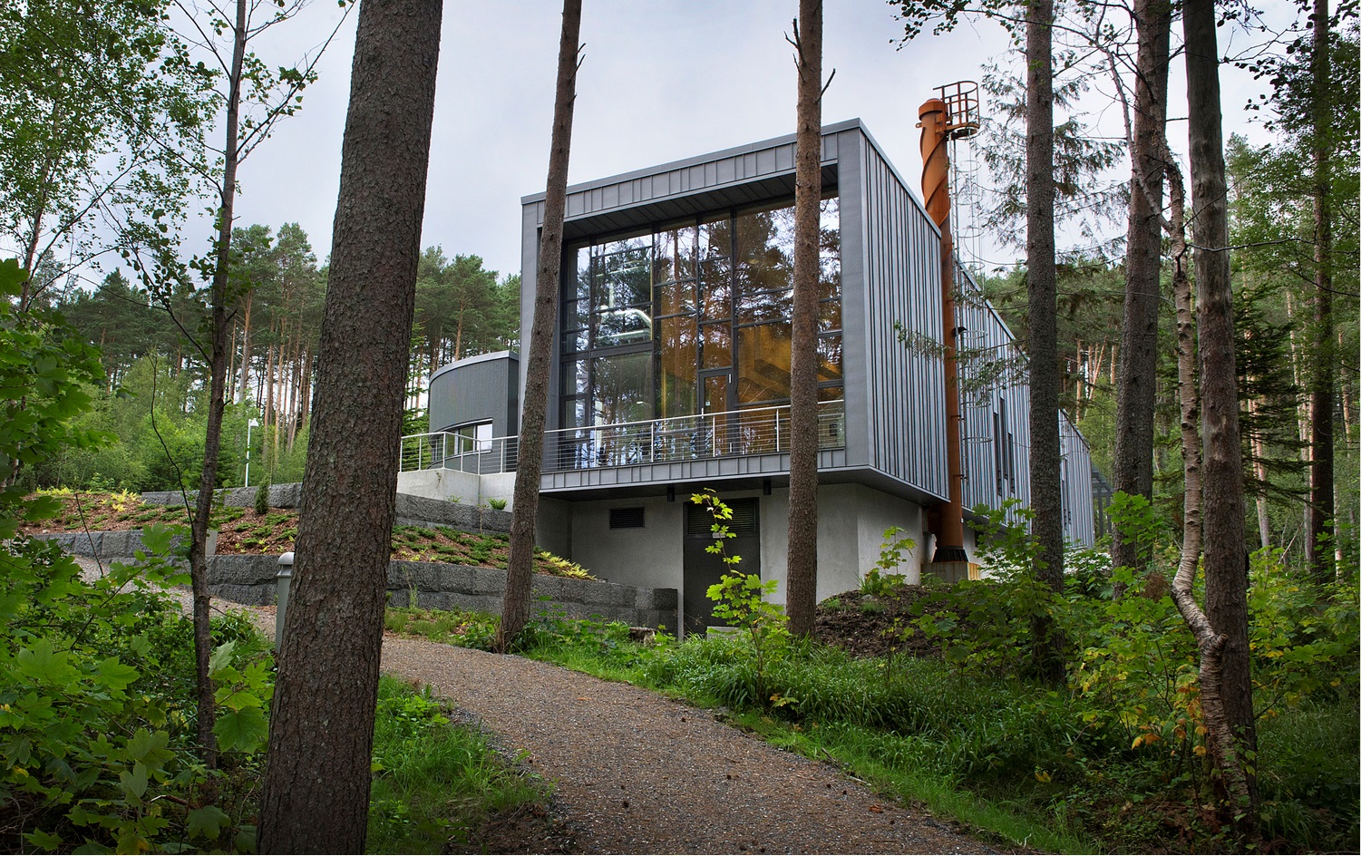 Ålesund krematorium, en moderne bygning med store glassflater og mørk metallkledning, omgitt av skog og en grussti som leder opp til inngangen. Foto.