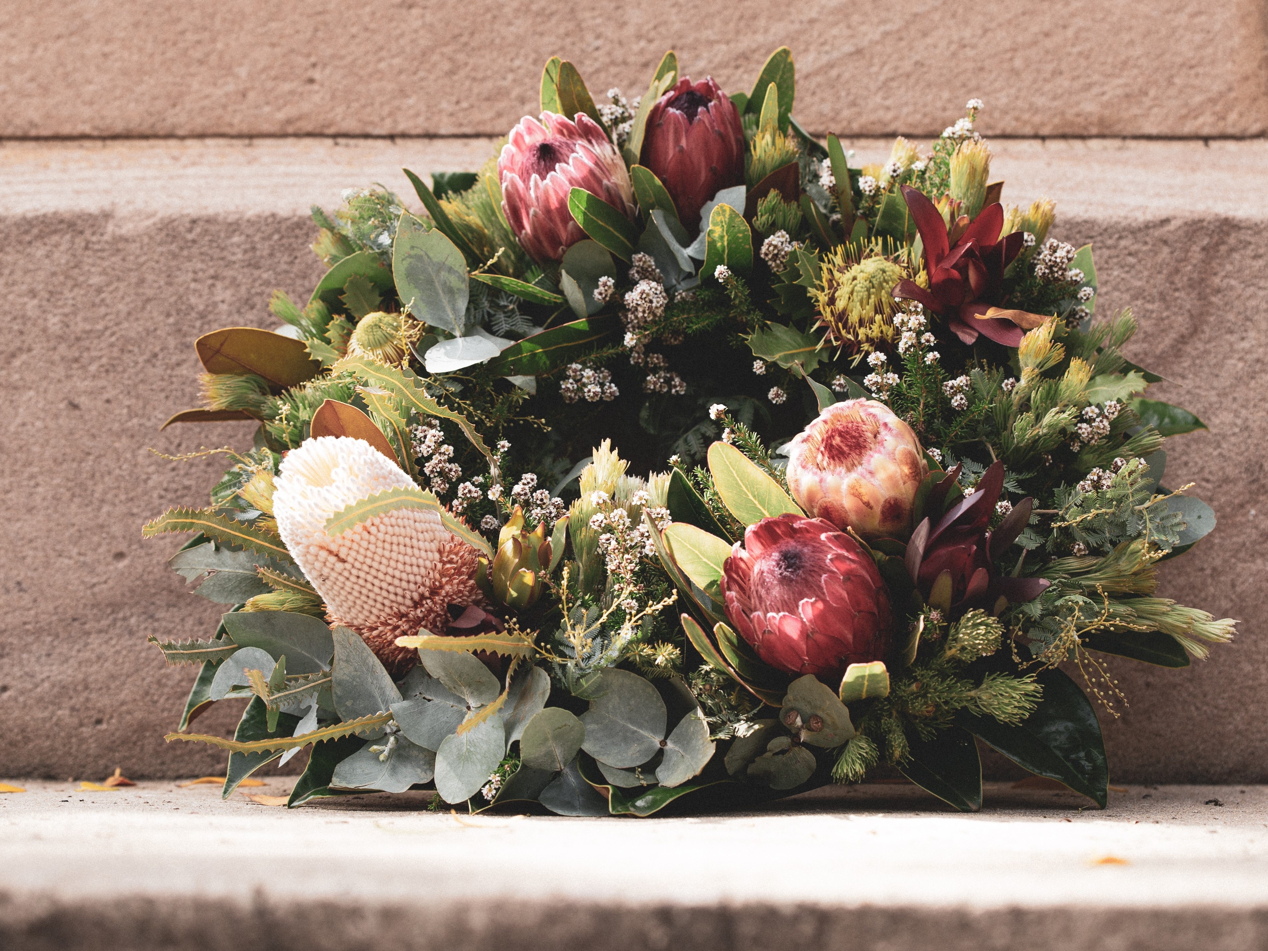 Blomsterkrans med røde protea og grønne blader hviler mot en trapp av lys stein. Foto.
