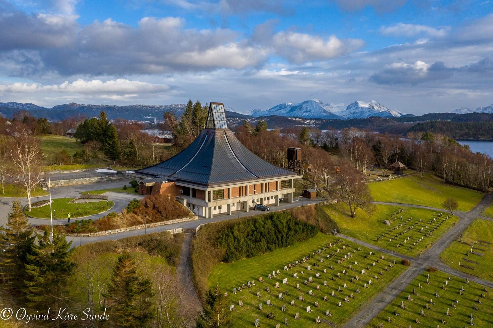 Dronebilde av en moderne kirke med skråtak og glasspyramide, omgitt av en gravplass. Fjorder, skog og snødekte fjell i bakgrunnen. Foto: Øyvind Kåre Sunde.