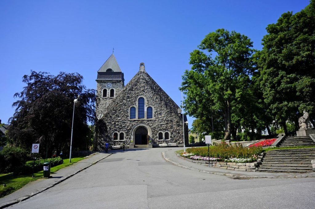 Steinkirke i jugendstil med et bratt skråtak og et firkantet klokketårn, omgitt av grøntområder og blomsterbed. En bred steintrapp fører opp til kirkeområdet. Blå himmel i bakgrunnen.