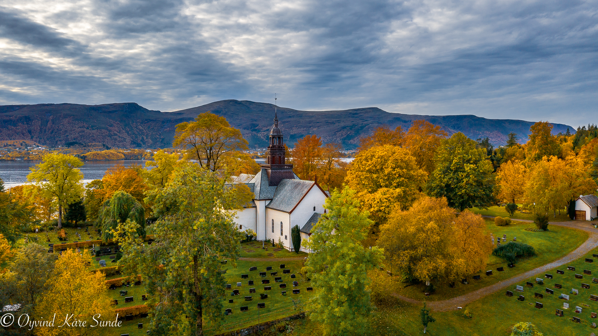 Hvit steinkirke med mørkt tak og rikt dekorert tårn, omgitt av høsttrær og fjordlandskap. Foto.