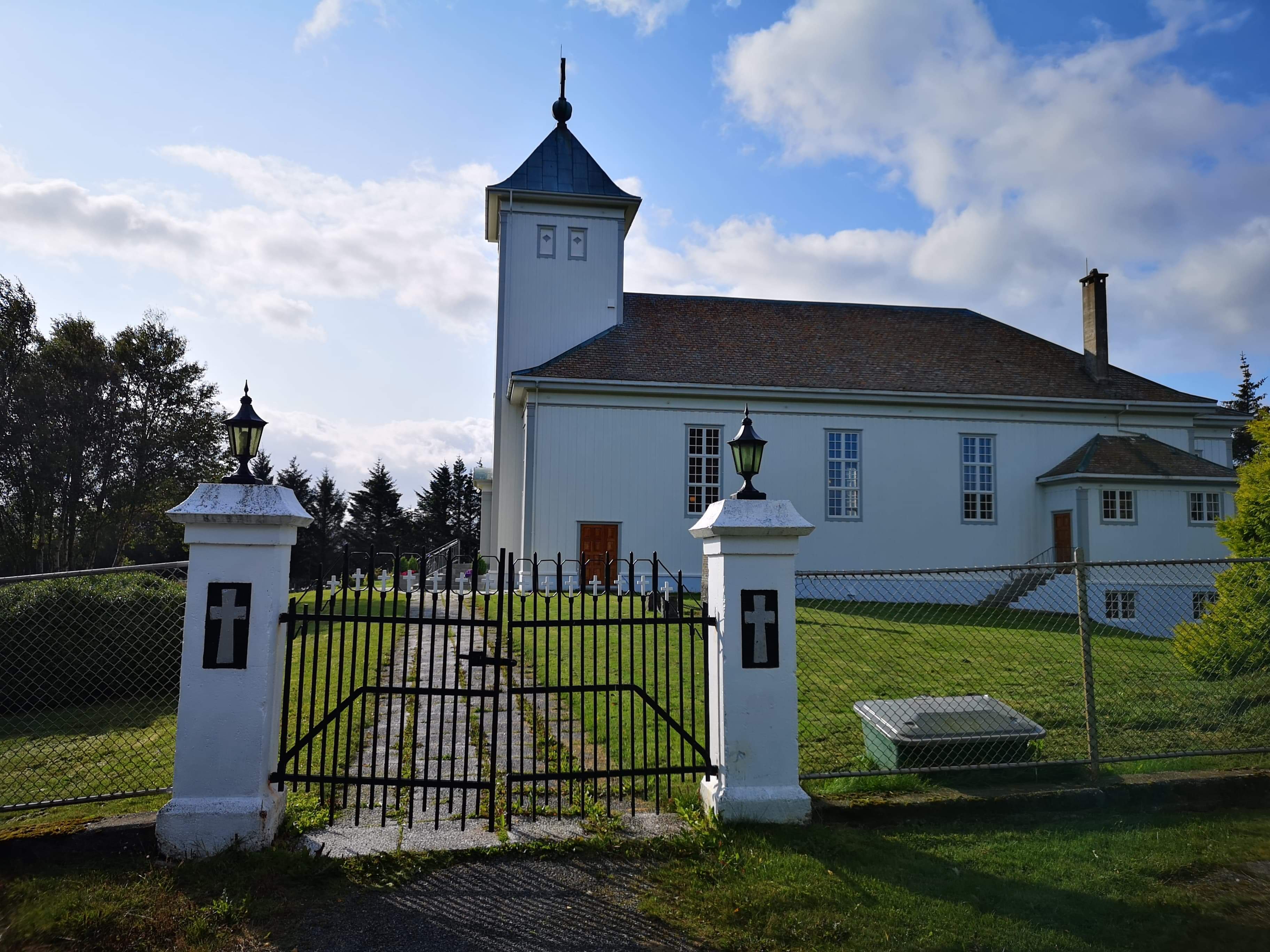Hvit trekirke med firkantet klokketårn, inngjerdet gravplass og smijernsport med kors. Foto.