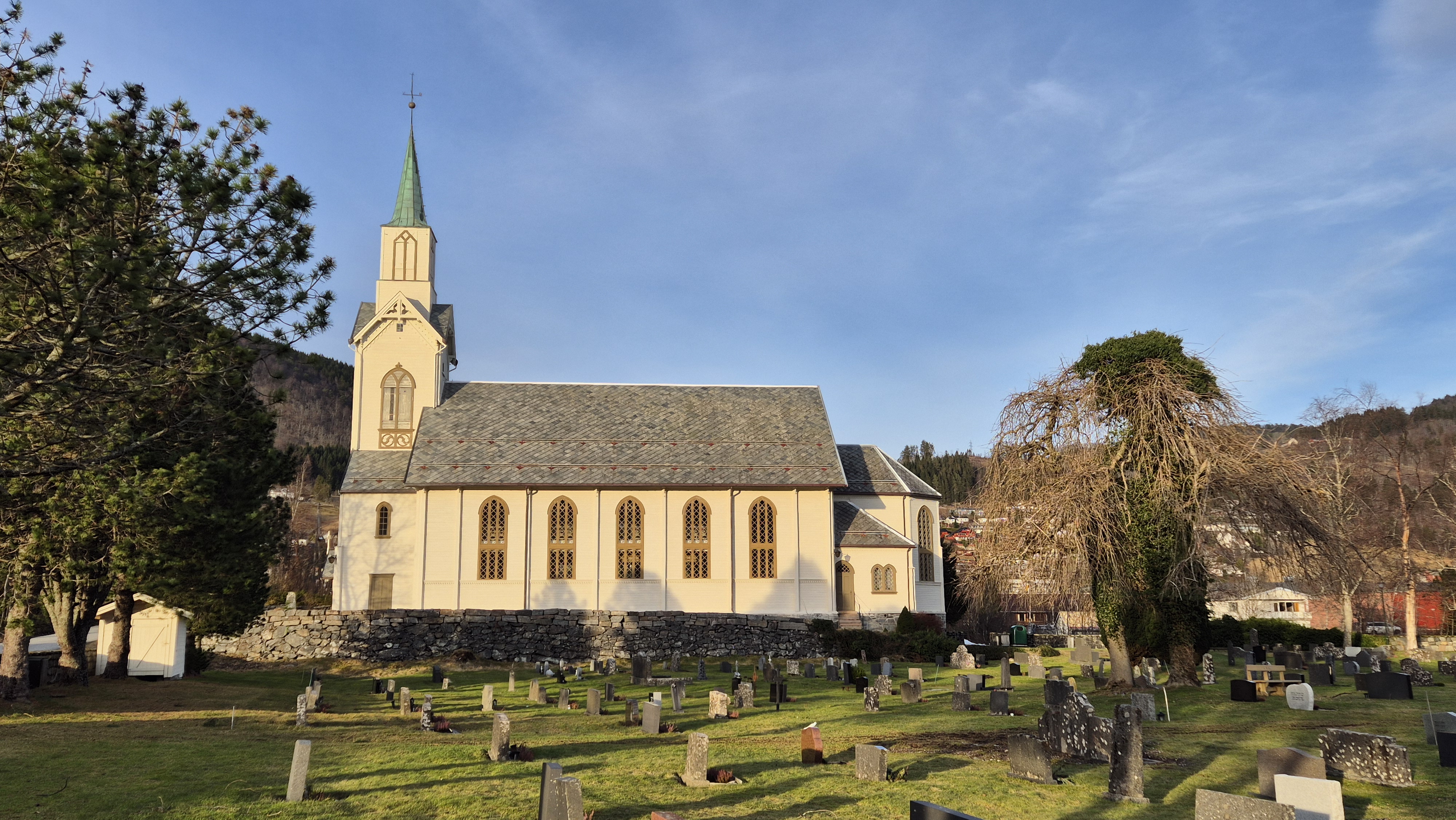 Lys trekirke med høye, smale vinduer og et grønt spir, omgitt av en gravplass og en gammel, mosegrodd steinmur. Landsbyen og åser sees i bakgrunnen under en blå himmel.