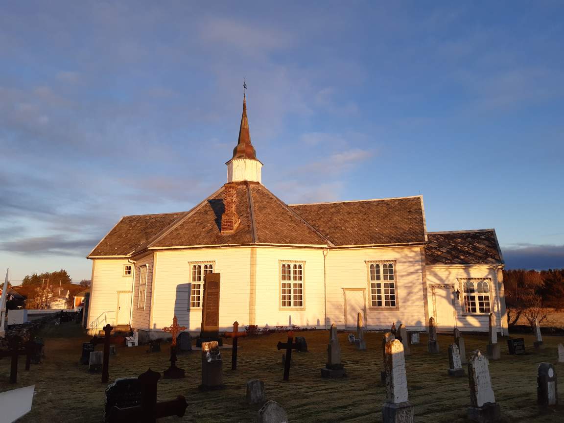 Et bilde av en hvitmalt trekirke med et spisst tårn og skifertak, badet i varmt sollys fra lav kveldssol. Kirken er omgitt av en gravplass med ulike gravstøtter og kors, og det er gress på bakken. I bakgrunnen ses trær, et åpent landskap og en delvis skyet himmel med blåtoner. Foto.