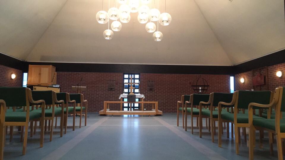 Kirkerom med mursteinsvegger, skråtak, rund lysekrone og grønnkledde stoler. Foto.