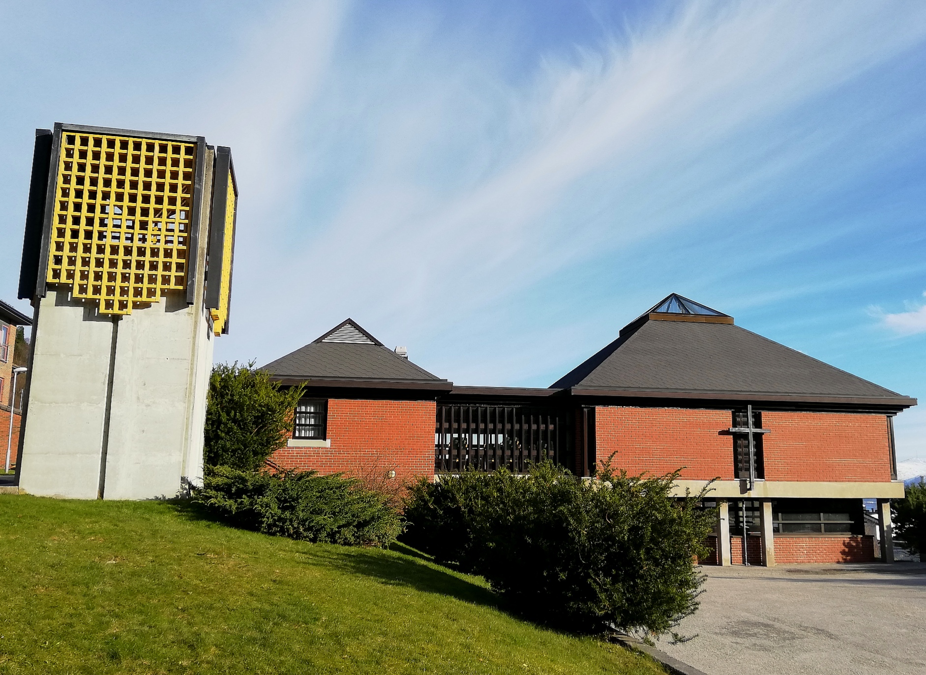Mursteinskirke med pyramideformede tak og betongklokketårn med gul gitterstruktur. Foto.