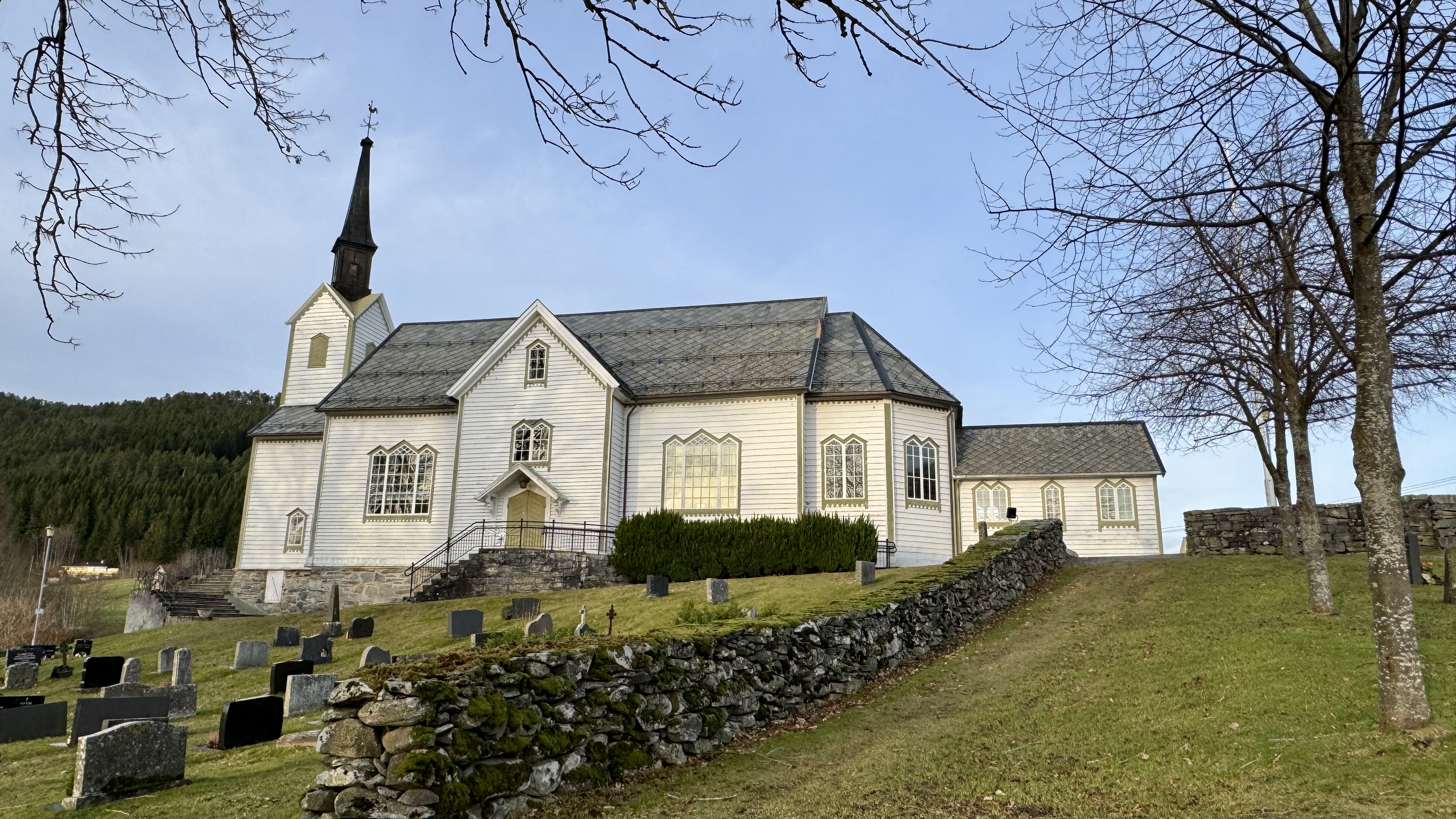 Hvit trekirke med bratt tak og slankt kirketårn, omgitt av en steinmur og gravplass. Kirken ligger i et grønt landskap med skog i bakgrunnen, under en klar himmel.
