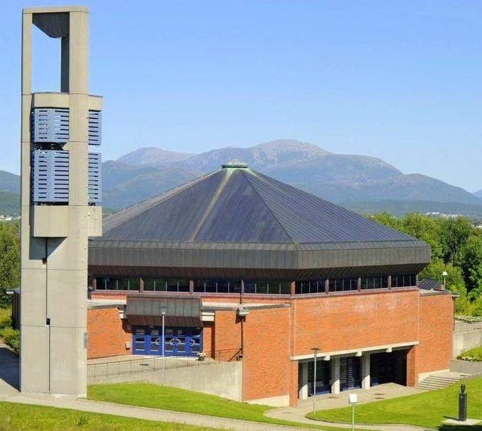 Moderne firkantet kirke i murstein med stort, mørkt pyramidetak og frittstående klokketårn. Foto.