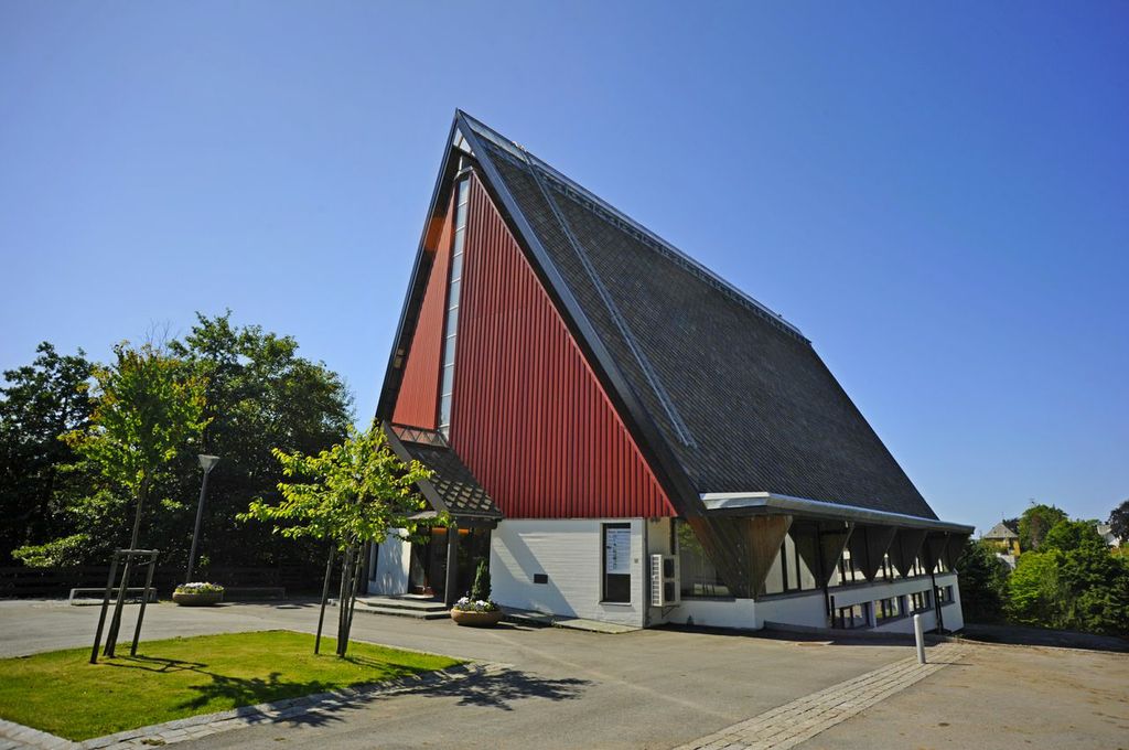 Moderne A-formet kirke med røde vegger, mørkt tak og høy, smal vindusrekke. Foto.