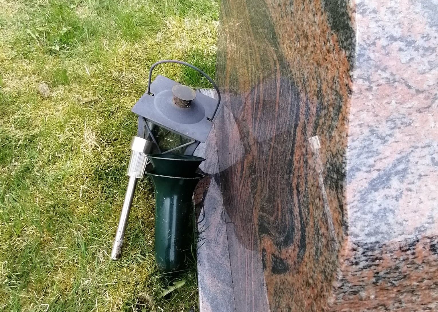 En lykt, en vase og en solcellelampe ligger inntil en gravstein på en gresskledd gravplass. Disse løse gjenstandene er plassert utenfor bedet, noe som kan være til hinder for daglig drift og vedlikehold av gravplassen. Foto.