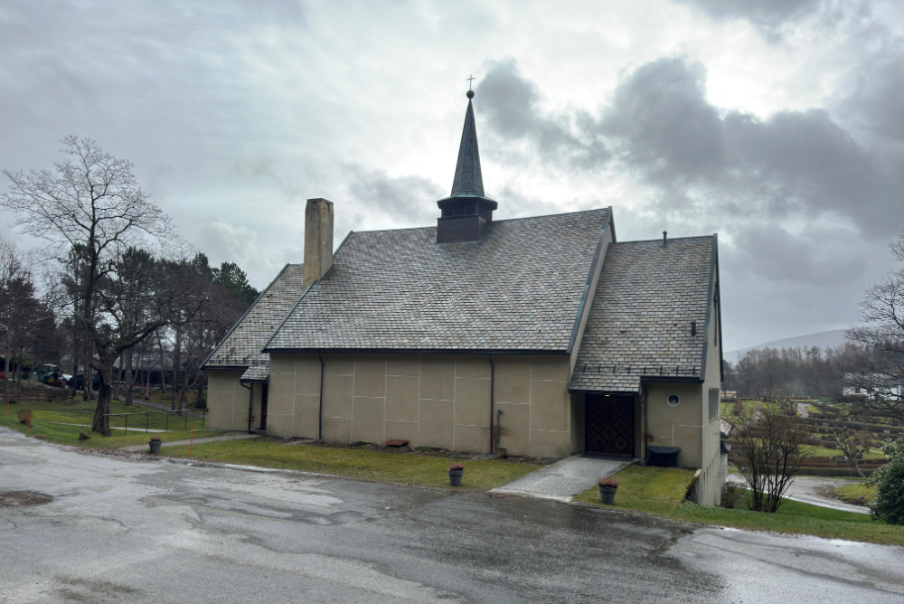 Et gammelt krematorium med beige fasade og skifertak under en overskyet himmel. Bygget har en karakteristisk spiss tårnspire og en liten inngangsdør til høyre. Området rundt er preget av vått asfaltdekke, gress, trær uten blader og en gravplass i bakgrunnen. Foto.